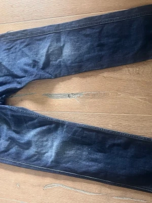 Mörkblå raka jeans - Säljer ett par mörkblå jeans med raka ben och diskreta slitningar. Jeansen har klassisk femficksdesign och kontrastsömmar. Materialet är denim i bomull och passformen är normal. Perfekta för en avslappnad och stilren look.