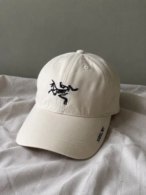 Arcteryx keps - Snygg vit keps från Arc'teryx med svart broderad logga framtill och text på skärmen. Klassisk böjd skärm och justerbar baksida. Tillverkad i bomull för en bekväm och avslappnad stil.