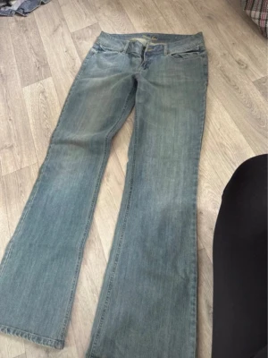 Blå bootcut jeans från Guess - Säljer ett par klassiska blå bootcut jeans från Guess med broderad logga på bakfickan. Jeansen har normal midja, fem fickor och är tillverkade i mjukt denimtyg. Perfekta för en avslappnad och trendig look.
