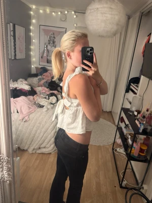 Vit ärmlös blus med volang och knyt - Supersöt vit blus med volangdetaljer på axlarna och knyt i ryggen. Toppen är ärmlös och har en luftig, croppad passform med v-ringad urringning. Perfekt för varma dagar och ger en romantisk vibe till din outfit.