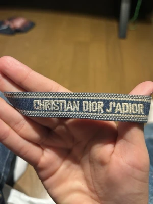 Christian Dior J'adior blått armband - Snyggt armband från Christian Dior med texten 'J'adior' broderat i vitt på blå textilband. Armbandet har justerbara knytband med tofsar i ändarna och en cool, avslappnad vibe. Perfekt accessoar för att lyfta din outfit med en touch av lyx.