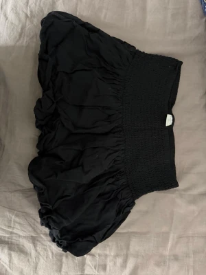 Svart kortkjol med resårmidja - Säljer en svart kortkjol med bred smockresår i midjan och puffig form. Kjolen har en luftig och voluminös siluett som ger en cool och avslappnad vibe. Perfekt att styla med en crop top eller hoodie.