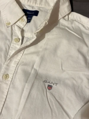 Vit skjorta från GANT med brodyr - Klassisk vit skjorta från GANT med broderad logga på bröstet. Skjortan har knappar framtill och på kragen, samt en stilren passform. Tillverkad i mjuk bomull som känns skön mot huden. Perfekt för dig som gillar en clean och tidlös stil.