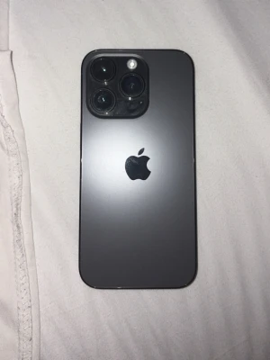 iPhone med svart baksida och trippelkamera - Snygg iPhone 14 pro med stilren svart baksida i glas och tre stora kameralinser. Telefonen har Apples logga i mitten och en modern, kantig design. Perfekt för dig som gillar att ta bilder och vill ha en mobil med premiumkänsla.