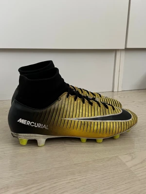 Nike Mercurial fotbollsskor guld/svart - Snygga Nike Mercurial fotbollsskor i guld och svart med vita detaljer. Skorna har högt skaft i strumpmodell, svarta snören och gula dobbar. Perfekta för dig som vill sticka ut på fotbollsplanen och se proffsig ut!