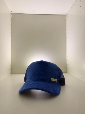 Blå truckerkeps från Maggiore - Snygg blå truckerkeps från Maggiore med böjd skärm och mesh på sidorna och bak. Kepsen har en mjuk, sammetsliknande framsida och justerbar rem bak. Märkesdetalj framtill och logotyp på insidan. Perfekt för en avslappnad streetwear-look.