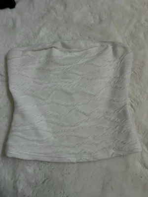 Vit bandeau topp i strukturerat tyg - Säljer en vit bandeau topp i ett strukturerat, lätt stretchigt tyg. Toppen är axelbandslös och har en figurnära passform som ger en clean och modern look. Perfekt att styla med jeans eller kjol för en trendig outfit.