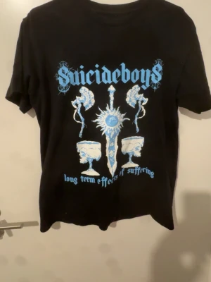  Suicideboys merch G59 - Säljer en svart t-shirt med stort blått och vitt Suicideboys-tryck fram och bak. Framsidan har text och grafiska motiv, baksidan har ett mönster av dödskallar i blått och vitt. Klassisk passform och rund hals. Perfekt för dig som gillar streetwear och bandmerch.