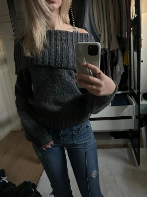 Mörkgrå off shoulder stickad tröja - Säljer en mörkgrå stickad tröja med bred ribbad krage som kan bäras off shoulder. Tröjan har en avslappnad passform, långa ärmar och ribbade muddar vid ärmslut och nederkant. Perfekt för kalla dagar och enkel att styla med jeans. Liten i storleken!