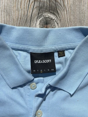 Ljusblå pikétröja från Lyle & Scott - Ljusblå pikétröja från Lyle & Scott med klassisk krage, knappslå och gul broderad logga på bröstet. Tillverkad i mjuk bomull och har korta ärmar för en clean och stilren look.