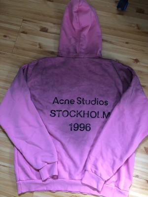 Rosa hoodie Acne Studios Stockholm - Säljer en rosa hoodie från Acne Studios med trycket 'Acne Studios STOCKHOLM 1996' på ryggen. Hoodien har huva och ribbade muddar vid ärmar och nederkant. Perfekt för dig som gillar streetwear och vill sticka ut med en färgstark look.