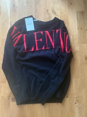  Valentino tröja - Snygg svart stickad tröja från Valentino med stor röd logotyp över bröstet och ärmen. Tröjan har rund halsringning och långa ärmar. Perfekt för dig som vill ha en trendig och bekväm look med exklusiv känsla.