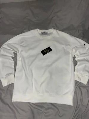 Vit sweatshirt från Stone Island - Säljer en clean vit sweatshirt från Stone Island med klassisk rund hals och ribbade muddar. Tröjan har den ikoniska logotypen på ärmen och är gjord i mjukt bomullsmaterial. Perfekt för dig som gillar stilrena plagg med streetwear-vibe.