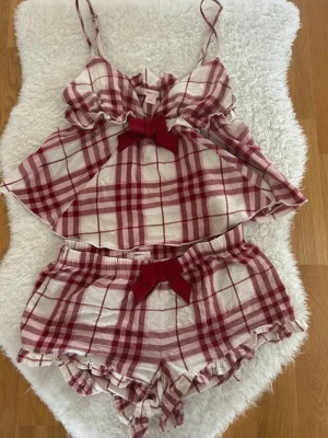 Rutig pyjamas-set med rosetter - Sött pyjamas-set med linne och shorts i röd- och vitrutigt mönster. Linnet har tunna axelband, volangkant och en röd rosett framtill. Shortsen har resår i midjan, volangkant och matchande röd rosett. Perfekt för mysiga kvällar.