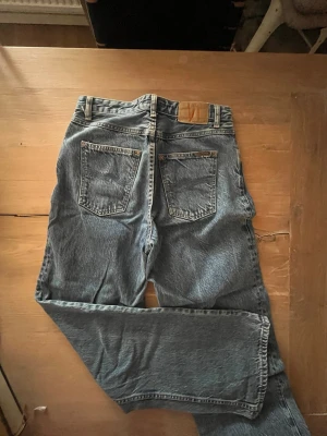 Blå raka jeans med hög midja - Säljer ett par klassiska blå jeans med rak passform och hög midja. Jeansen har två bakfickor och en läderpatch i midjan. Tillverkade i slitstarkt denimtyg, perfekta för en avslappnad och tidlös look.