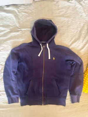 Marinblå hoodie från Polo Ralph Lauren - Säljer en marinblå hoodie från Polo Ralph Lauren med dragkedja, vit dragsko och klassisk gul logga på bröstet. Hoodien har känguruficka och ribbade muddar. Tillverkad i mjuk bomull för skön känsla. Perfekt för en avslappnad och stilren look. Priset är inte hugget i sten så skriv om de är något. 