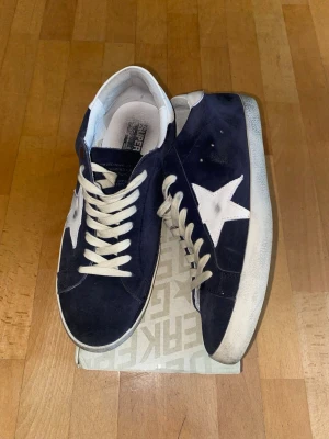 Mörkblå golden goose sneakers - Snygga mörkblå sneakers i mocka med vit stjärndetalj på sidan och vita skosnören. Skorna har en klassisk låg profil och vit sula. Perfekta för dig som gillar streetwear och vill ha något som sticker ut lite extra.