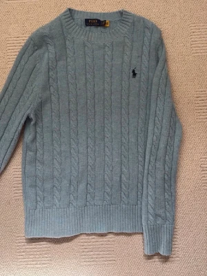 Ljusblå kabelstickad tröja Ralph Lauren - Kabelstickad tröja i ljusblå från Polo Ralph Lauren. Klassisk rund halsringning, ribbade muddar och nederkant samt den ikoniska broderade loggan på bröstet.