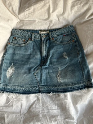 Blå jeanskjol med slitningar - Säljer en blå jeanskjol med slitna detaljer och rå kant nertill. Kjolen har klassiska fem fickor och knappgylf. Från märket Tally Weijl