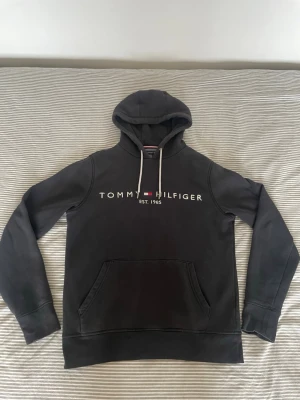 Svart hoodie Tommy Hilfiger - Snygg svart hoodie från Tommy Hilfiger med vit logga och text på bröstet samt EST. 1985. Klassisk känguruficka framtill och vita dragsnören i huvan. Tillverkad i mjukt bomullsmaterial och har ribbade muddar vid ärmar och nederkant.