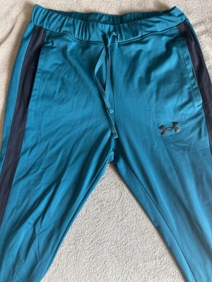 Blå mjukisbyxor från Under Armour - Snygga blå mjukisbyxor från Under Armour med svarta sidopaneler och dragsko i midjan. Tillverkade i mjukt och stretchigt material som passar perfekt för chill eller träning. Logga på vänster ben för extra sportig vibe.