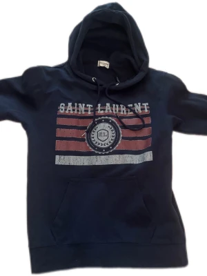 Saint Laurent hoodie - Mörkblå hoodie med huva och dragsko, stor magficka och tryck framtill med texten 'Saint Laurent' samt rödvita ränder och emblem. Mjuk bomullskänsla och avslappnad passform, perfekt för en chill look.