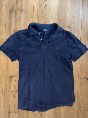Marinblå pikétröja från Polo Ralph Lauren - Klassisk marinblå pikétröja från Polo Ralph Lauren med korta ärmar och två knappar vid kragen. Tröjan har den ikoniska röda logotypen broderad på bröstet och är tillverkad i bomull för en bekväm känsla.    Nypris:1085kr.     Mitt pris:599kr