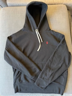 Ralph Lauren Hoodie - Säljer en svart hoodie från Polo Ralph Lauren med klassisk röd logga broderad på bröstet. Hoodien har vit dragsko, känguruficka och är gjord i mjukt bomullsmaterial. Perfekt för en chill och stilren look. Storlek M, passar även S