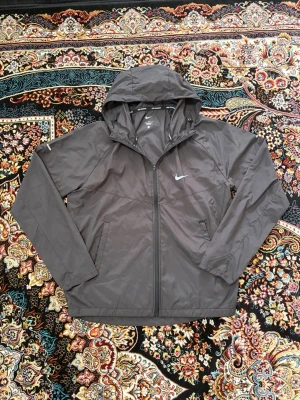 Svart vindjacka från Nike Running - Snygg svart vindjacka från Nike Running med huva och dragkedja framtill. Jackan har två sidofickor och är tillverkad i lätt, vindtätt material. Perfekt för löpning eller andra utomhusaktiviteter. Diskret Nike-logga på bröstet och tryck på insidan.