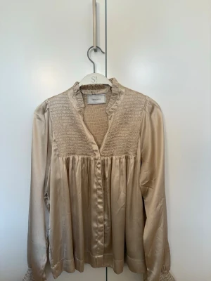 Beige glansig blus med smockdetaljer - Snygg beige blus i glansigt material med smock upptill och volangkantad krage. Blusen har v-ringning och långa ärmar med breda manschetter. Perfekt för dig som gillar en chic och trendig look.