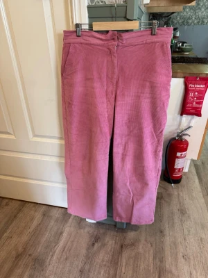 Rosa raka manchesterbyxor - Rosa manchesterbyxor från Indiska med raka ben och klassisk ribbad struktur. Modellen har elastisk midja, knapp- och dragkedjestängning samt praktiska sidfickor. Ett bekvämt och stilrent plagg i bomull med stretch – perfekt för både vardag och avslappnade tillfällen.  Material: 97% Bomull, 3% Elastan  Benets innerlängd: 74 cm  Röd blus från Kappahl med romantiska detaljer som en stor volang över axlarna, liten ståkrage med knytband samt långa ärmar med elastiska volangmuddar för en feminin och e