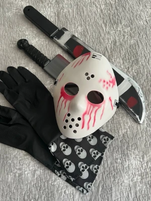 Jason Voorhees maskeradkit - Maskeradkit med vit hockeymask med röda bloddetaljer, svarta handskar, svart bandana med dödskalletryck samt två plastvapen – en machete och en kniv. Perfekt för skräcktema eller Halloween.
