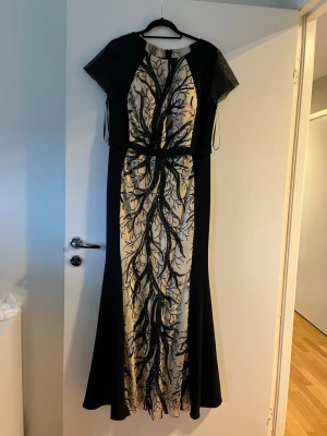 Svart långklänning med broderi - Elegant svart långklänning med korta ärmar och vackert broderat mönster i svart på beige botten framtill. Klänningen har en figurnära passform upptill och faller snyggt nedtill. Perfekt för speciella tillfällen.