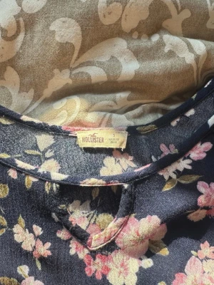 Blommig blus från Hollister - Mörkblå blus från Hollister med rosa och vita blommor. Blusen har smala axelband, en liten öppning vid halsen och lång ärm med offshoulder-design. Materialet känns mjukt och luftigt, perfekt för en trendig look.