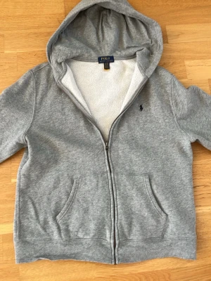 Grå hoodie med dragkedja från Polo - Säljer en grå hoodie från Polo med dragkedja framtill och två stora fickor. Insidan är mjuk och fleecefodrad, perfekt för extra komfort. Klassisk huva och diskret Polo-logga broderad på bröstet. Materialet är bomullsmix som känns riktigt skön mot huden. Står xl på lappen men passade min brorsa som har xs-s