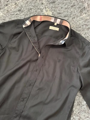 Svart skjorta från Burberry - Snygg svart skjorta från Burberry med klassiskt rutigt mönster på insidan av kragen och manschetterna. Skjortan har långa ärmar, knappar framtill och diskret broderad logga på bröstet. Perfekt för dig som gillar stilrena detaljer.