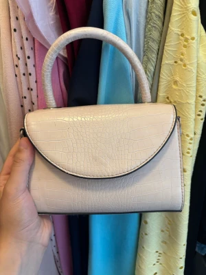 Ljusbeige handväska med krokoprägling - Snygg ljusbeige handväska i konstläder med krokodilpräglad yta och rundat lock. Väskan har ett stadigt handtag och silverfärgade metalldetaljer på sidorna. Perfekt accessoar för att lyfta din outfit.