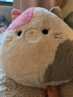 Stor mjuk katt Squishmallow - Supermjuk och rund Squishmallow i form av en katt med beige, grå och rosa detaljer. Katten har broderat ansikte, små öron och svans samt fluffig plyschyta. Perfekt som kudde eller gosedjur för mysiga stunder.💕