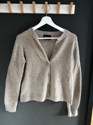 Beige stickad kofta från Pieces - Mysig beige stickad kofta från Pieces med knappar framtill. Koftan har lång ärm och ribbade muddar vid ärmslut och nederkant. Perfekt att slänga över en topp för en avslappnad look.