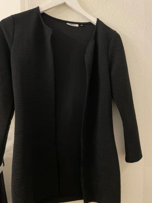 Svart ribbad cardigan jacka från ONLY - Svart långärmad cardigan jacka från ONLY i storlek XS. Jackan har en öppen front utan knappar eller dragkedja och är tillverkad i ett ribbat syntetmaterial. Perfekt för lager-på-lager och enkel att matcha med olika outfits.