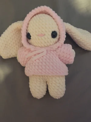 Virkad kanin i rosa hoodie - Söt handgjord kanin i mjukt garn med stora öron och svartvita ögon. Kaninen har en ljusrosa hoodie med luva och snören, och kroppen är vit. Perfekt som dekoration eller gosedjur.