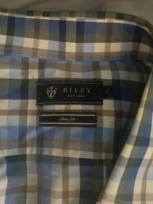 Rutig skjorta från Riley slim fit - Säljer en rutig skjorta från Riley i slim fit-modell. Skjortan har blå, gröna, bruna och vita rutor, långa ärmar och knappar framtill. Perfekt för dig som gillar klassisk stil med modern passform.