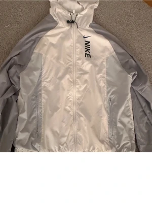 Vit och grå vindjacka från Nike - Snygg vit och ljusgrå vindjacka från Nike med dragkedja framtill, huva med dragsko och två fickor med dragkedja. Jackan har en sportig look och är tillverkad i lätt, glansigt material som passar perfekt för blåsiga dagar.