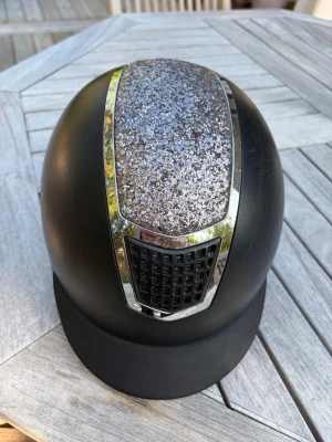 Svart ridhjälm med glitterdetalj CRW 56-58 - Svart ridhjälm från CRW med glittrig panel upptill och silvriga detaljer. Justerbart hakspänne och vadderad insida för skön passform.