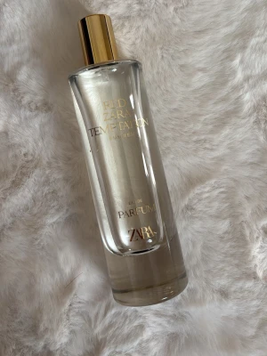 ZARA Red Temptation Eau de Parfum - ZARA Red Temptation Eau de Parfum i en elegant, genomskinlig glasflaska med guldfärgat lock. Flaskan har en stilren och modern design med guldtext på framsidan. Perfekt för dig som gillar lyxiga och fräscha dofter. 80ml ungefär nu efter användning 
