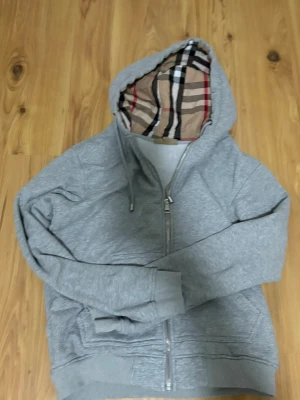 Grå hoodie med rutigt foder - Snygg grå hoodie med dragkedja och huva. Insidan av huvan och öppningen har ett klassiskt beige, svart och rött rutigt mönster. Hoodien har snörning i huvan och ribbade muddar vid ärmar och nederkant. Perfekt för en chill och stilren look.