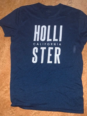 Blå Hollister t-shirt med tryck - Snygg blå t-shirt från Hollister med stort vitt tryck på bröstet där det står 'Hollister California'. Klassisk rund hals och korta ärmar. Perfekt för en avslappnad och trendig stil.