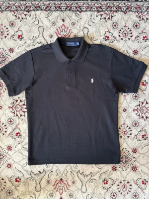 Svart pikétröja från Polo Ralph Lauren - Klassisk svart pikétröja från Polo Ralph Lauren med vit broderad logga på bröstet. Tröjan har korta ärmar, krage och knappar vid halsen. Perfekt till jeans eller shorts för en clean och stilren look.