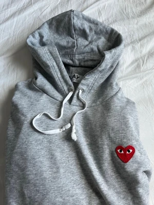Play hoodie - Snygg grå hoodie med huva och vita dragsnören. På bröstet sitter ett broderat rött hjärta med ögon, vilket ger en unik och lekfull touch. Hoodien har fickor på sidorna och är tillverkad i mjukt sweatshirtmaterial. Perfekt för en avslappnad och trendig stil.