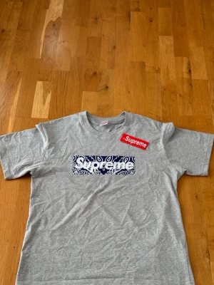 Grå Supreme t-shirt med paisleymönster - Säljer en grå t-shirt från Supreme med klassisk box logo framtill i blått och vitt paisleymönster. T-shirten har korta ärmar och normal passform. Perfekt för dig som gillar streetwear och vill sticka ut med en ikonisk design.
